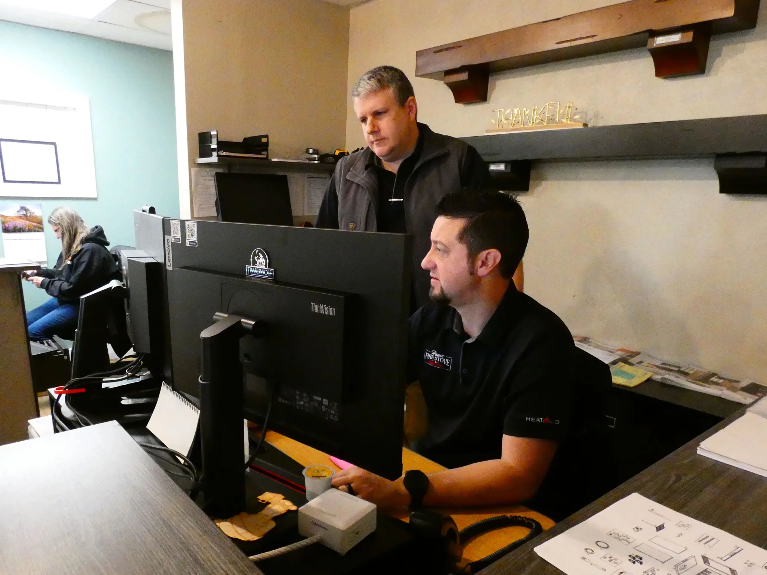 Two men working on desktop computers in an office with shelves and paperwork around. A woman in the background is using a phone and laptop. Thankful sign is on a shelf.