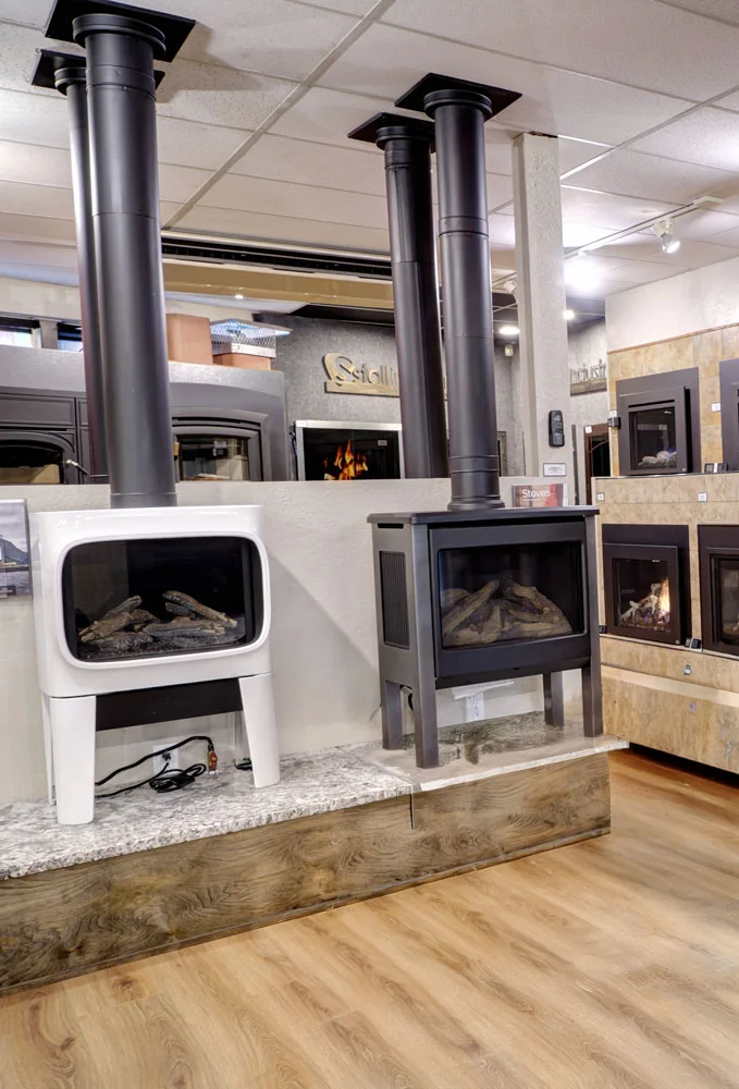 Showroom with modern wood stoves and fireplaces. Two stoves, one white, one black, are on display with long black chimneys extending to the ceiling. Other models are visible in the background.