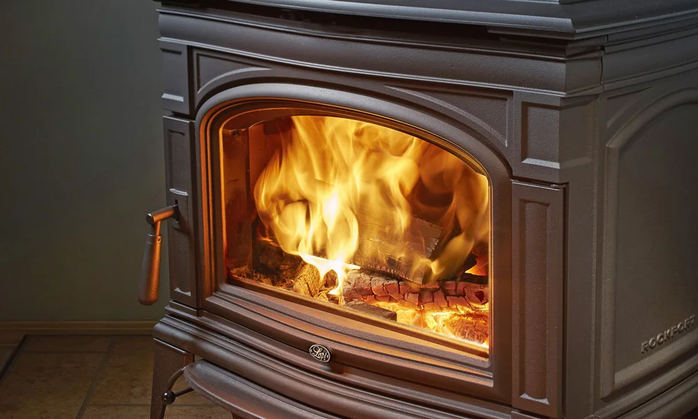 A black cast iron wood burning stove with firewood inside that is actively burning, casting an orange glow through the glass door on the front. There is a handle on the left side.