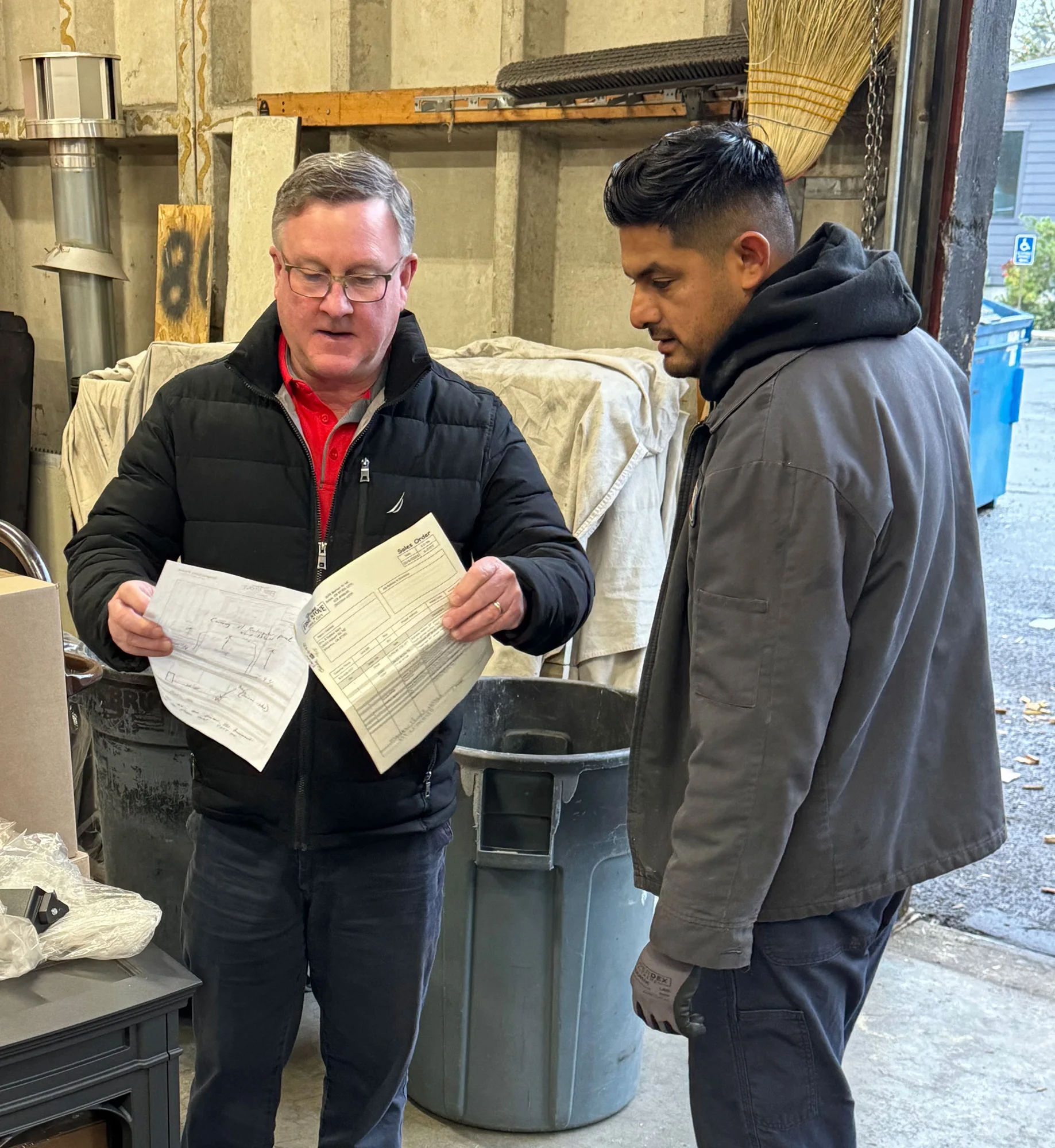 Two men standing in a workshop. One man, dressed in a black jacket, is holding and showing documents to the other man, who is dressed in a gray jacket.