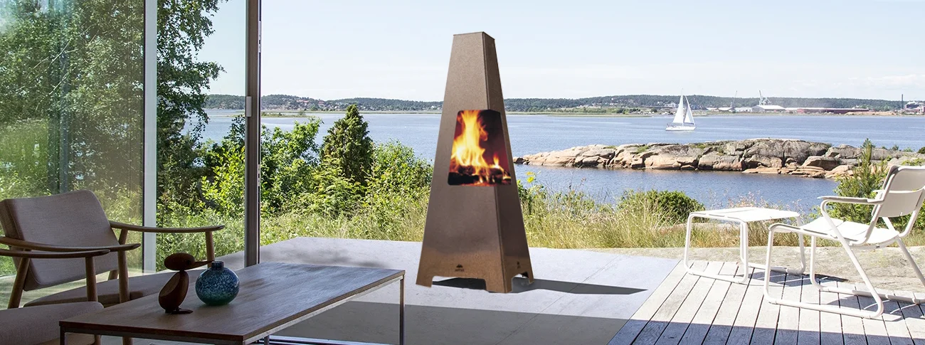 Outdoor patio with a tall metal fireplace, two chairs, and a table. The patio overlooks a scenic lake with a boat sailing and rocky shoreline in the background.
