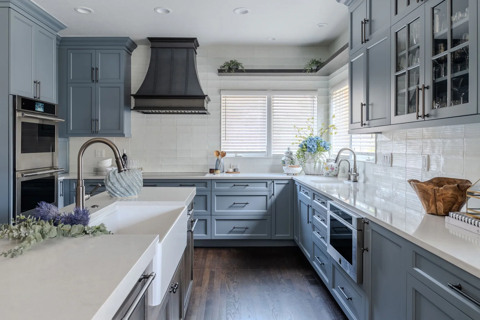 Modern kitchen with blue cabinets, white countertops, and stainless steel appliances. A farmhouse sink, decorative plants, and wooden accents complete the design.