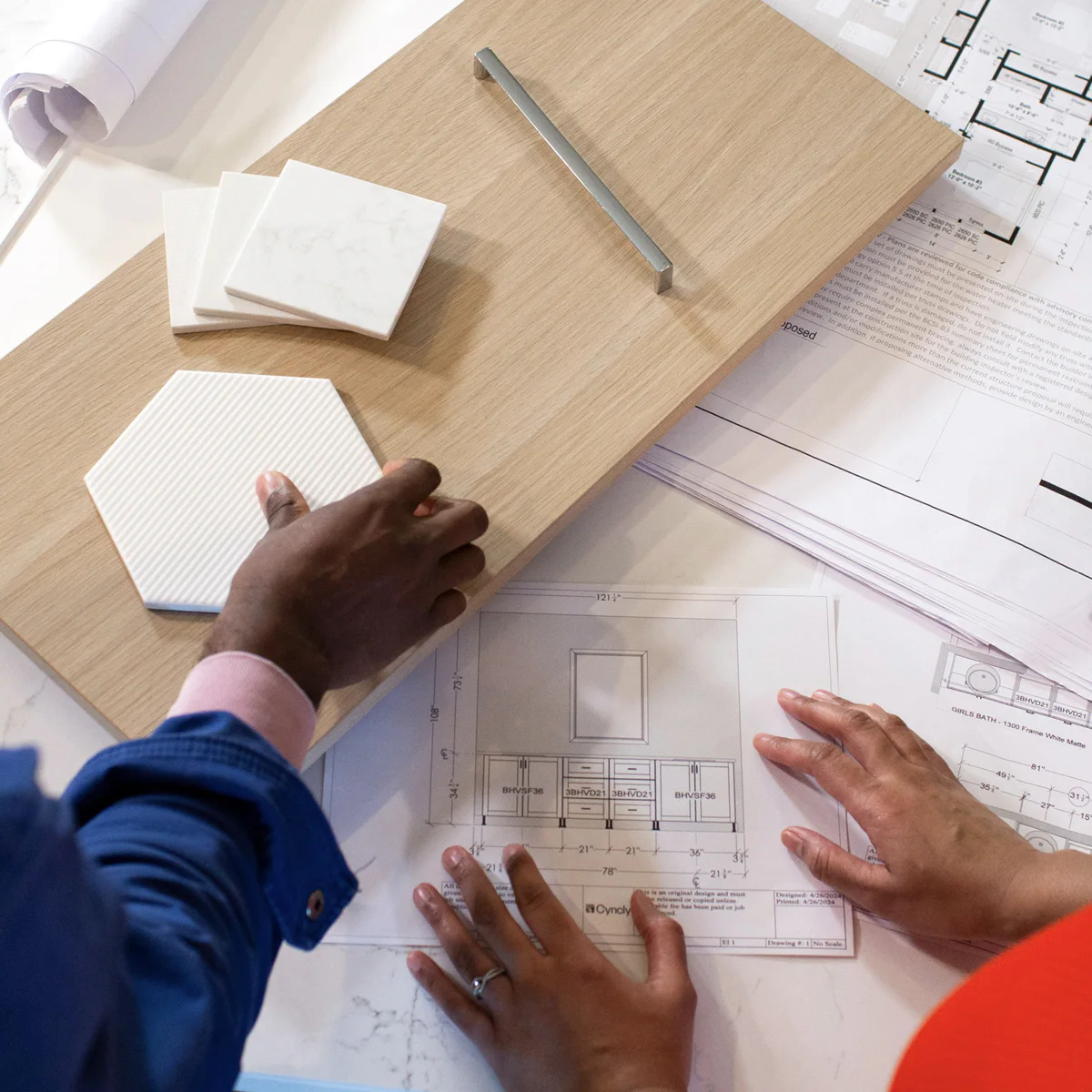 A wooden board with a metal handle, hexagonal and square tiles on top, and hands examining architectural blueprints, indicating an interior design planning session.