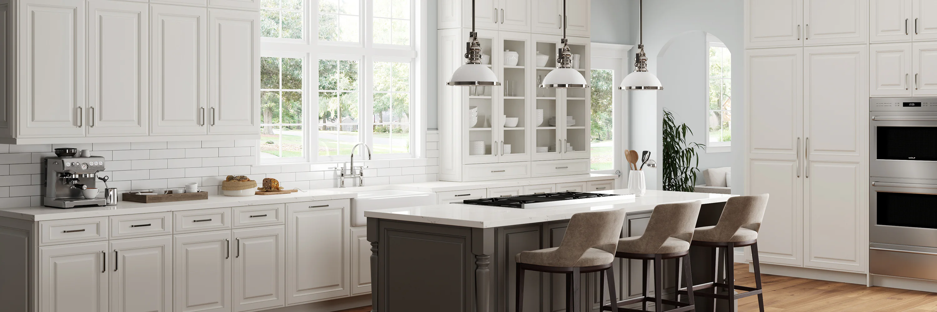 Modern kitchen with white cabinets, a large island with beige bar stools, pendant lights, and stainless steel appliances. A coffee maker and tray sit on the counter. Large windows provide natural light.