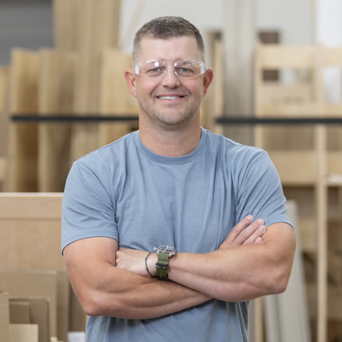 Man in a gray t-shirt and safety glasses stands with arms crossed in a workshop. He is smiling, and wood materials are blurred in the background.