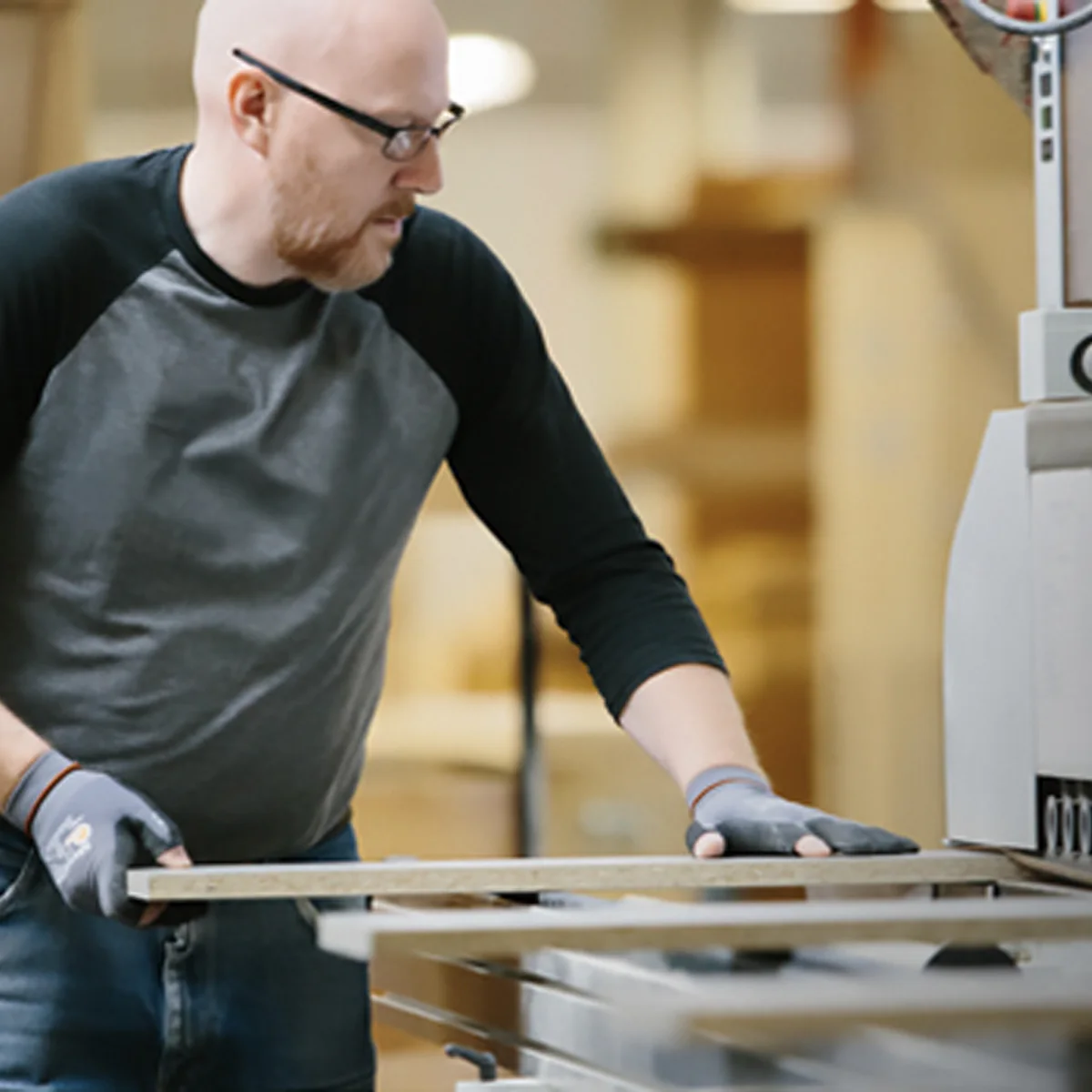 Man in safety glasses and gloves operates a woodworking machine, carefully feeding a wooden board through the equipment in a workshop setting.