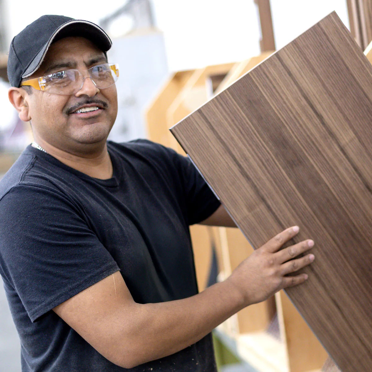 Man in safety glasses and a cap holding a wooden panel in a workshop, smiling.