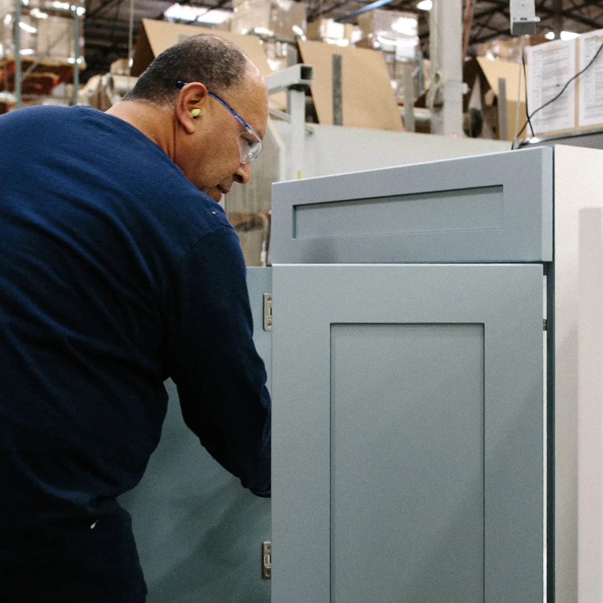 A person wearing ear protection and safety glasses, assembling blue industrial cabinets in a factory setting with machinery and packaging materials in the background.