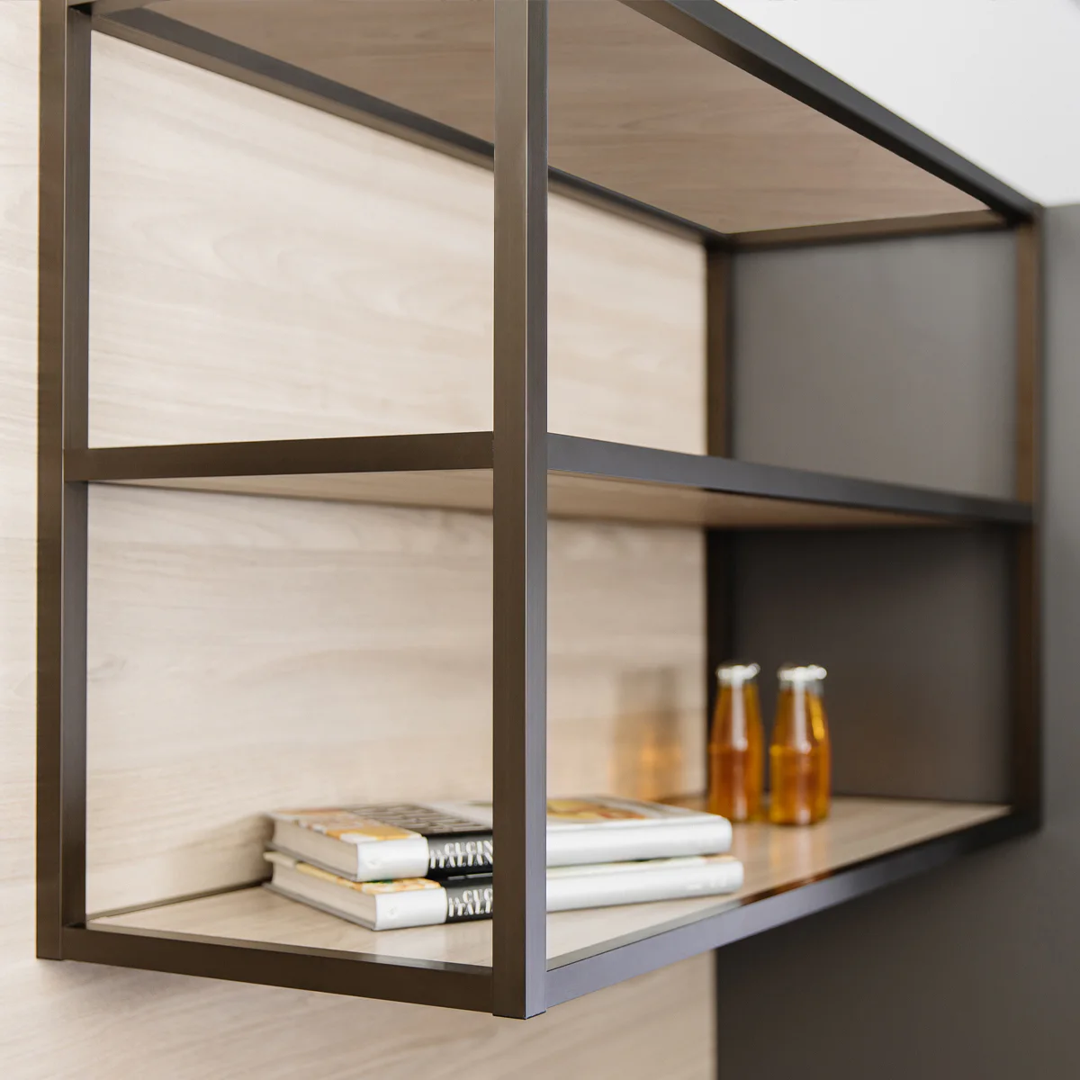 Modern shelving unit with a dark metal frame and wooden shelves, displaying several closed books and three small glass bottles with orange liquid on a light wood background.