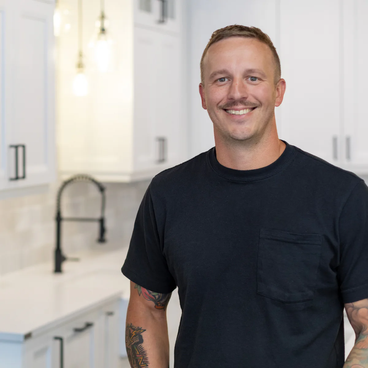 A person with tattoos smiles while standing in a modern kitchen with white cabinets, black handles, and a black faucet under bright lights.