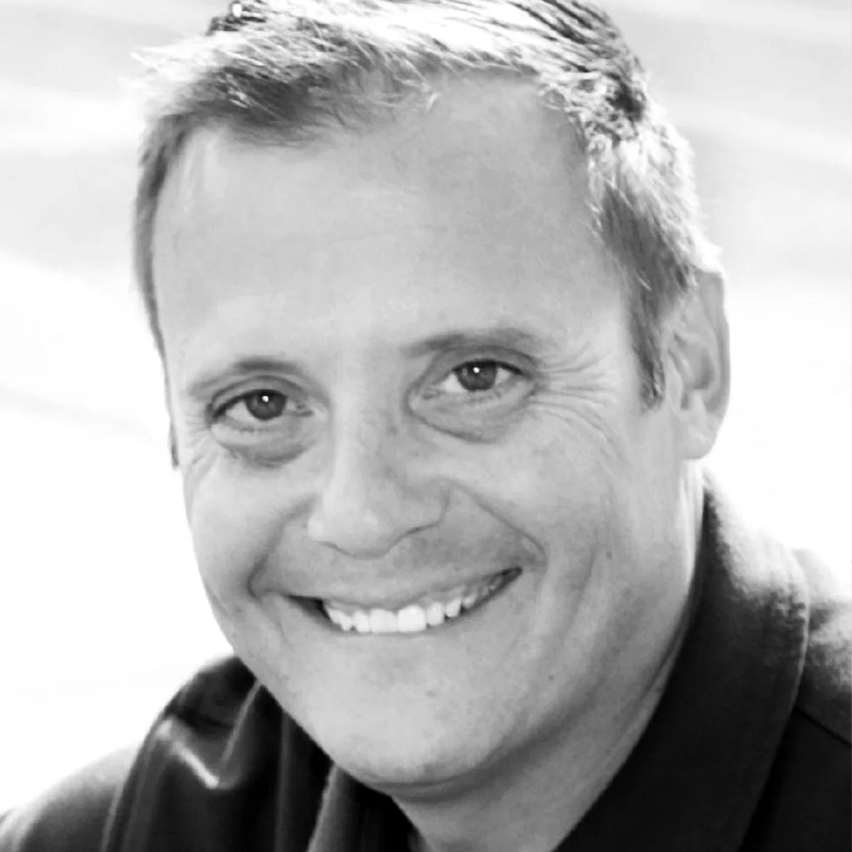 Smiling man with short hair in a black and white close-up portrait, wearing a dark shirt, looking directly at the camera.