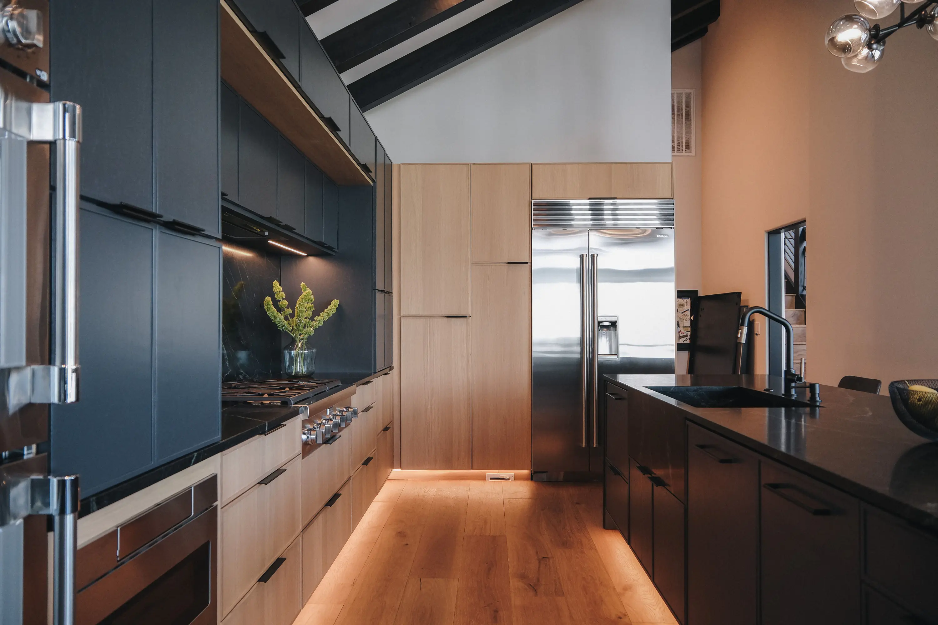 Modern kitchen with dark cabinets, wood accents, stainless steel appliances, and under-cabinet lighting. A vase with green flowers sits on the counter, and a sleek faucet is visible.