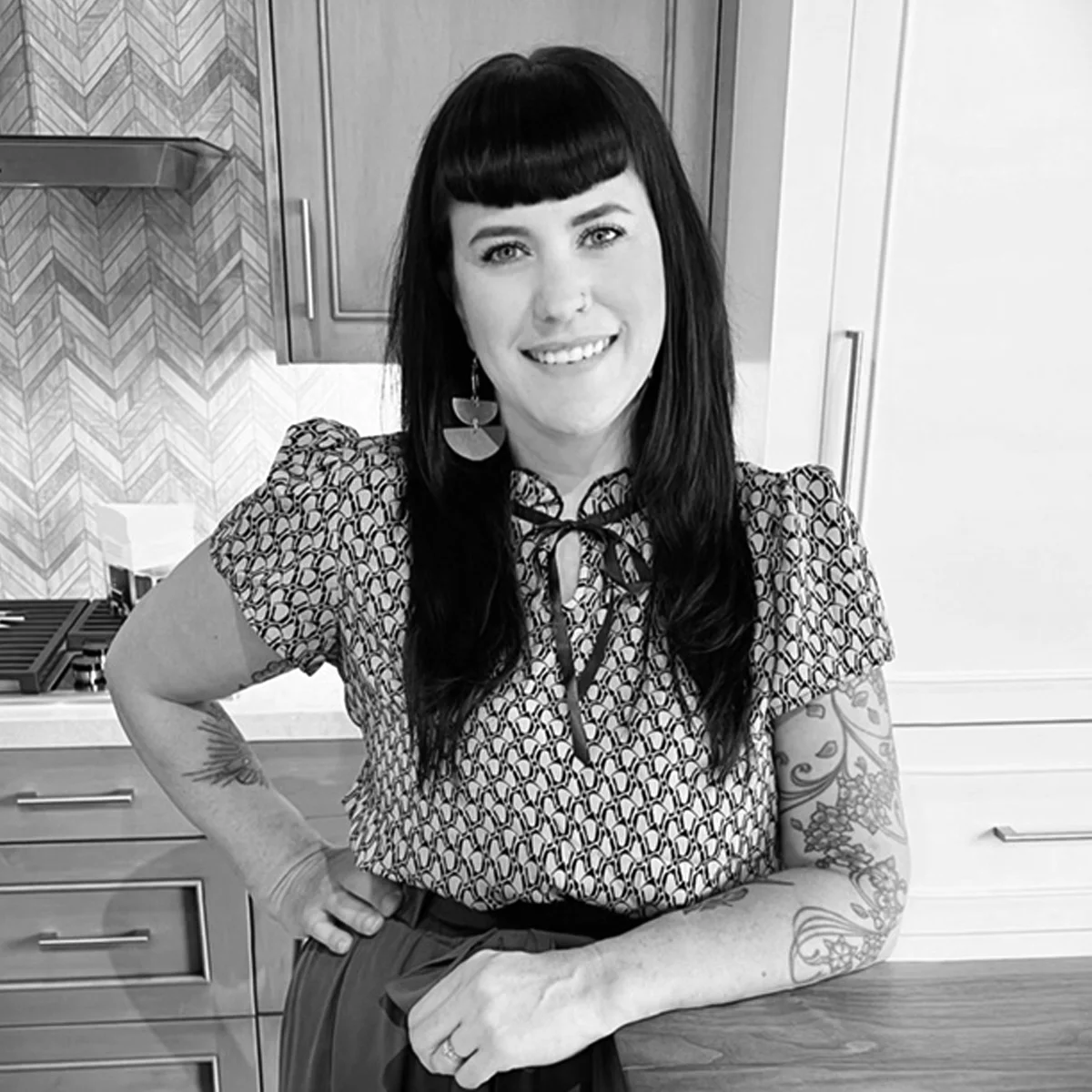 Smiling woman with long dark hair stands in a kitchen, wearing a patterned blouse and statement earrings. She has tattoos on her left arm and rests her hand on a counter.