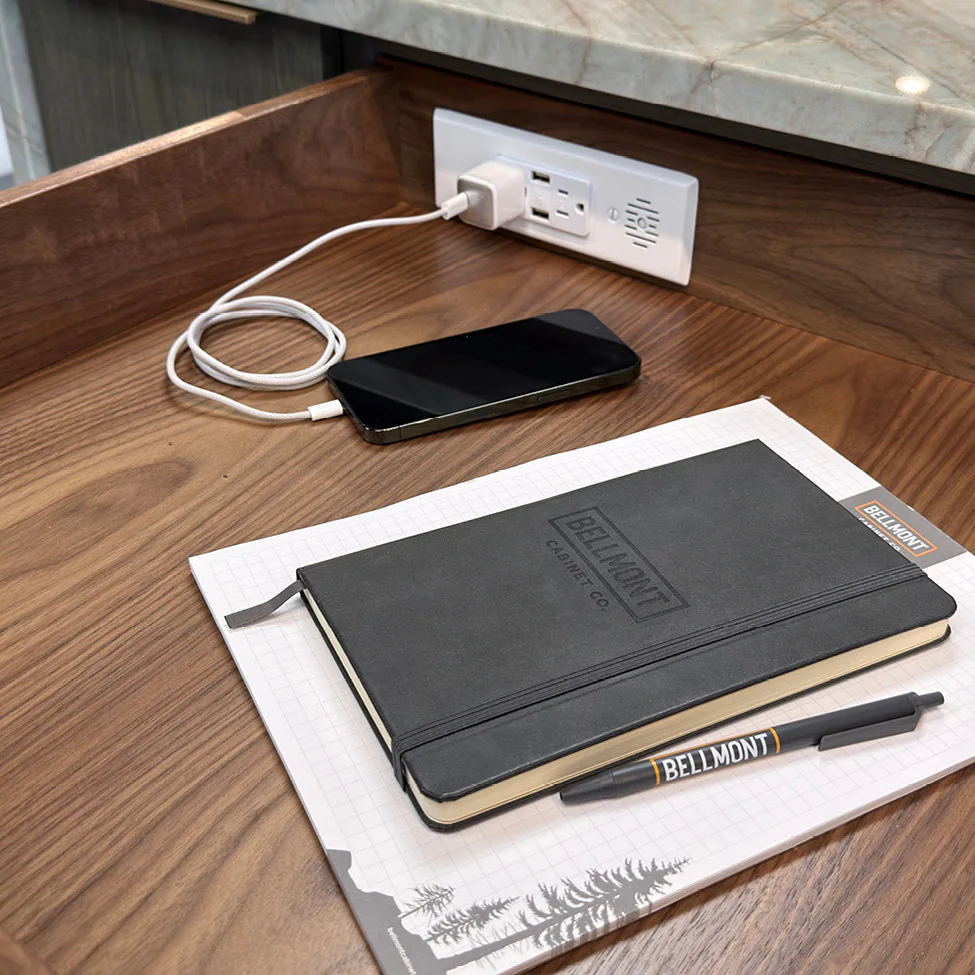 Black Bellmont notebook and pen on graph notepad beside a charging smartphone plugged into a wall outlet on a wooden desk