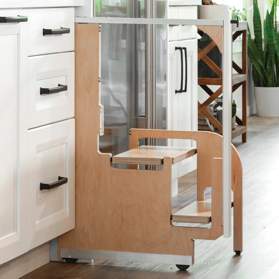 Wooden pull-out kitchen step stool (learning tower) beside white cabinets with black handles.