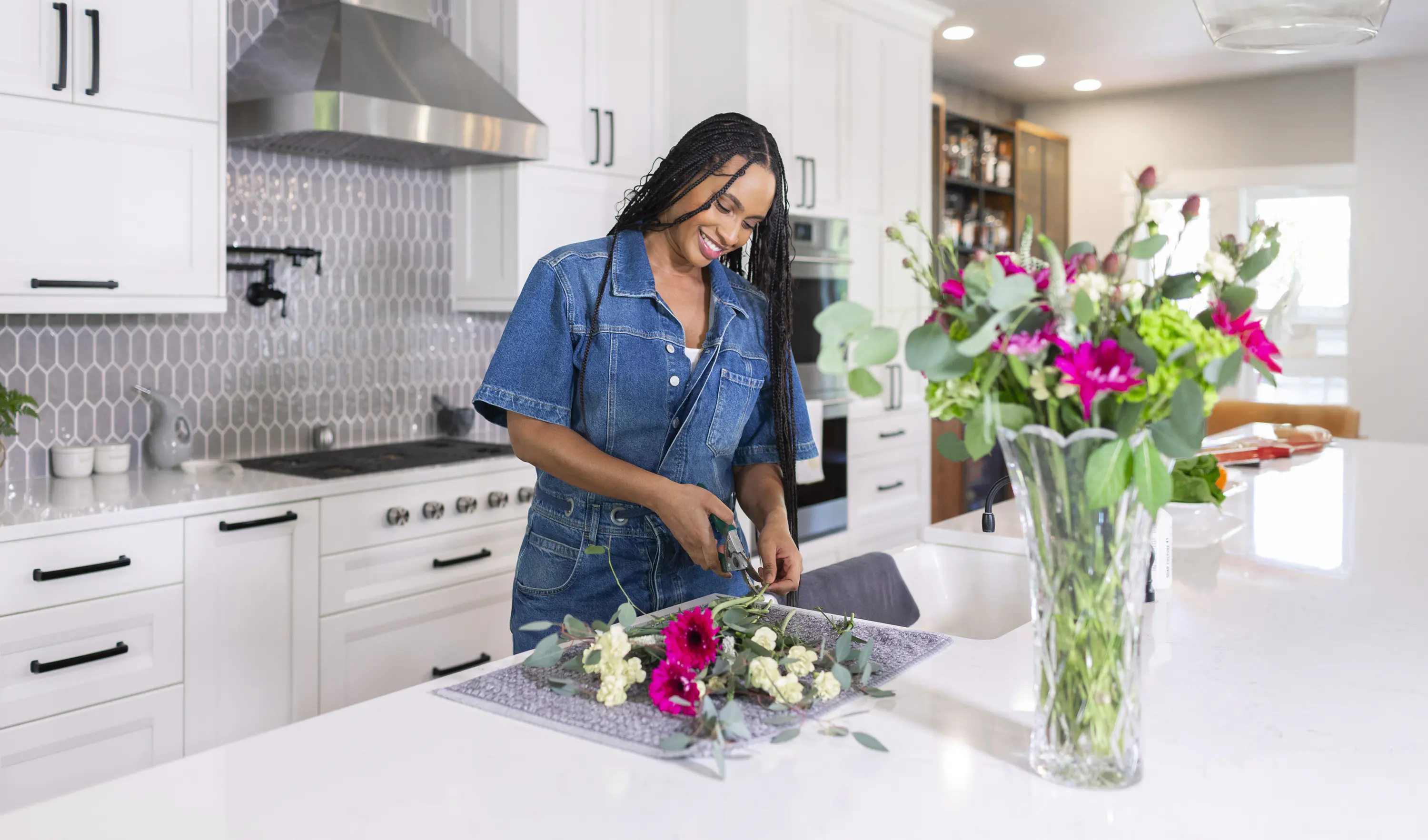 Woman in a denim jumpsuit smiles while arranging a bouquet of pink and white flowers on a kitchen counter with white cabinets and a vase in the foreground.
