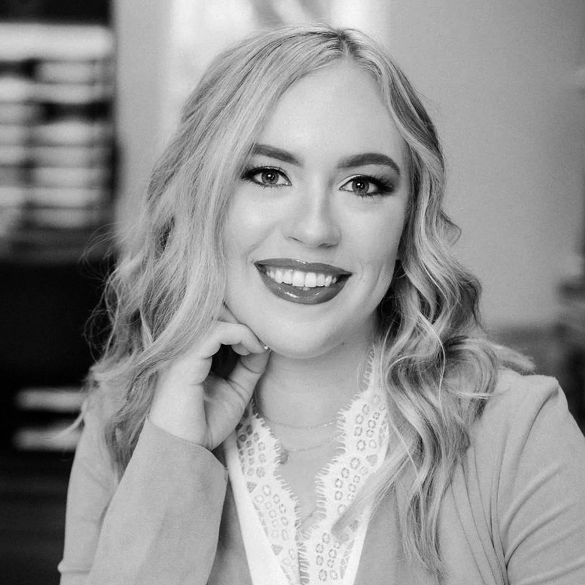 Black and white portrait of a smiling person with long, wavy hair, resting their chin on their hand. They're wearing a light-colored blazer and a patterned blouse.