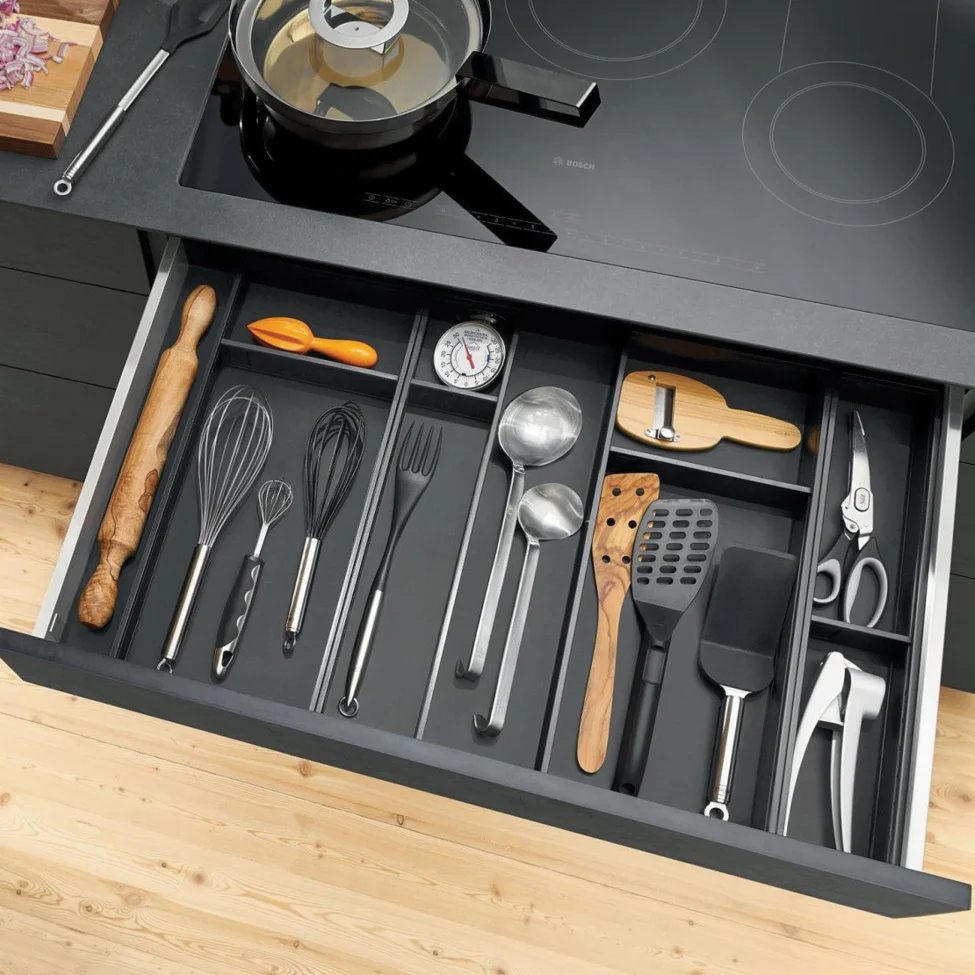Open kitchen drawer under stovetop with neatly organized cooking utensils: whisks, ladles, spatulas, rolling pin, scissors.