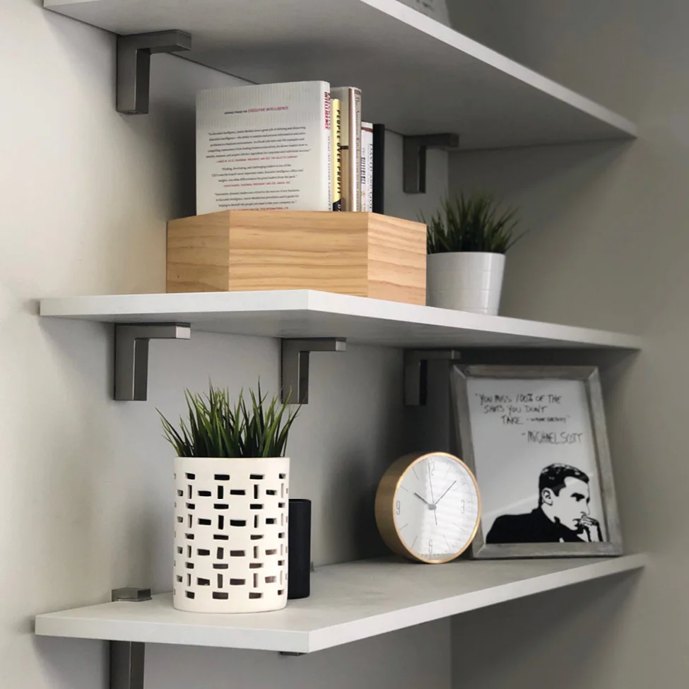 White floating shelves holding a wooden box, books, two potted plants, a round clock, and a framed black-and-white portrait.