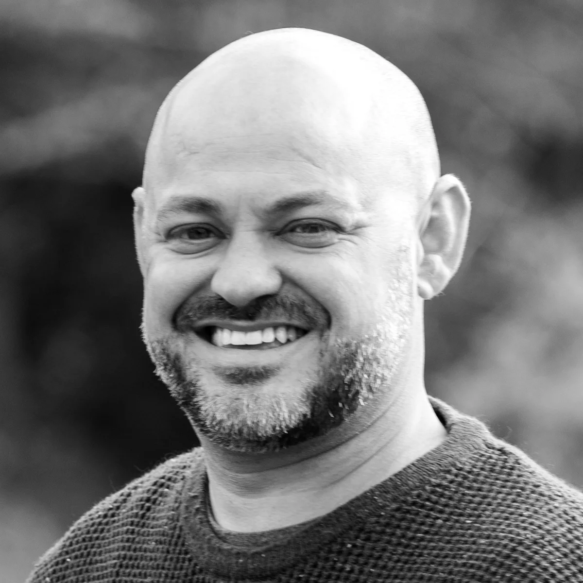 A black and white portrait of a bald man with a beard, smiling broadly while wearing a textured sweater. The background is blurred, creating a soft focus effect.