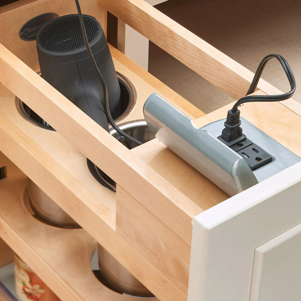 Wooden drawer organizer with circular canister slots and a black appliance cord plugged into a pop-up power outlet.