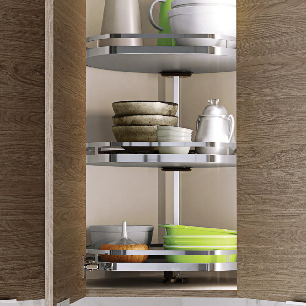 Open wood kitchen cabinet with three-tier chrome lazy Susan holding stacked bowls, a silver teapot, and green bowls.