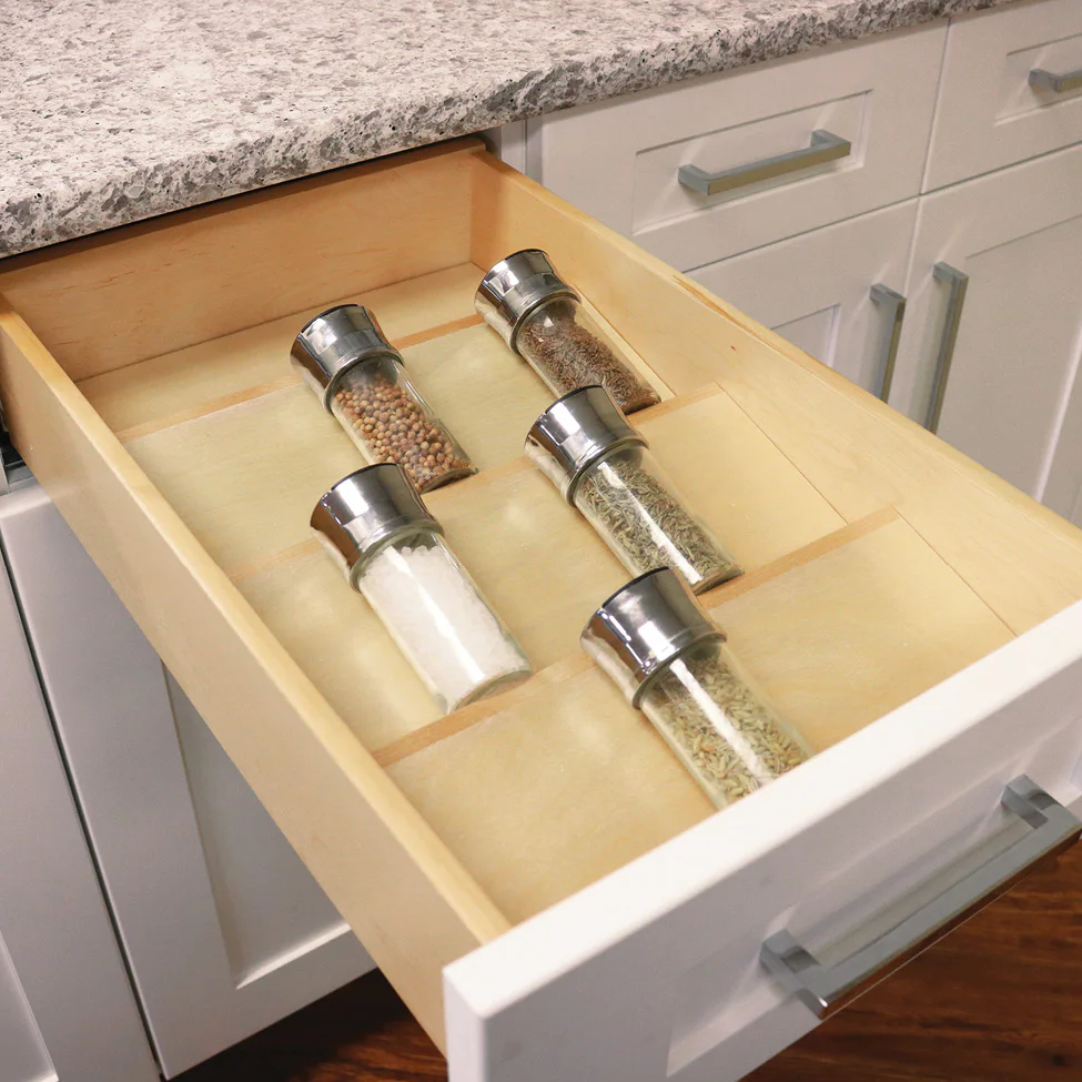 Open kitchen drawer with six glass spice jars in wooden dividers beneath a granite countertop