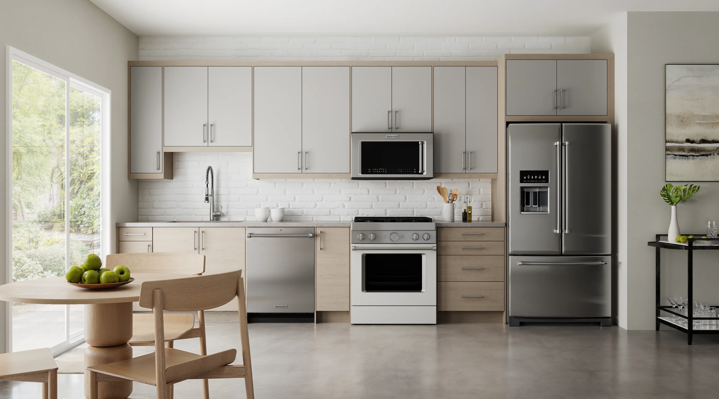 Modern kitchen with light wood and gray cabinets, a stainless steel fridge, oven, and dishwasher. A round table with green apples and a sliding glass door to the left.