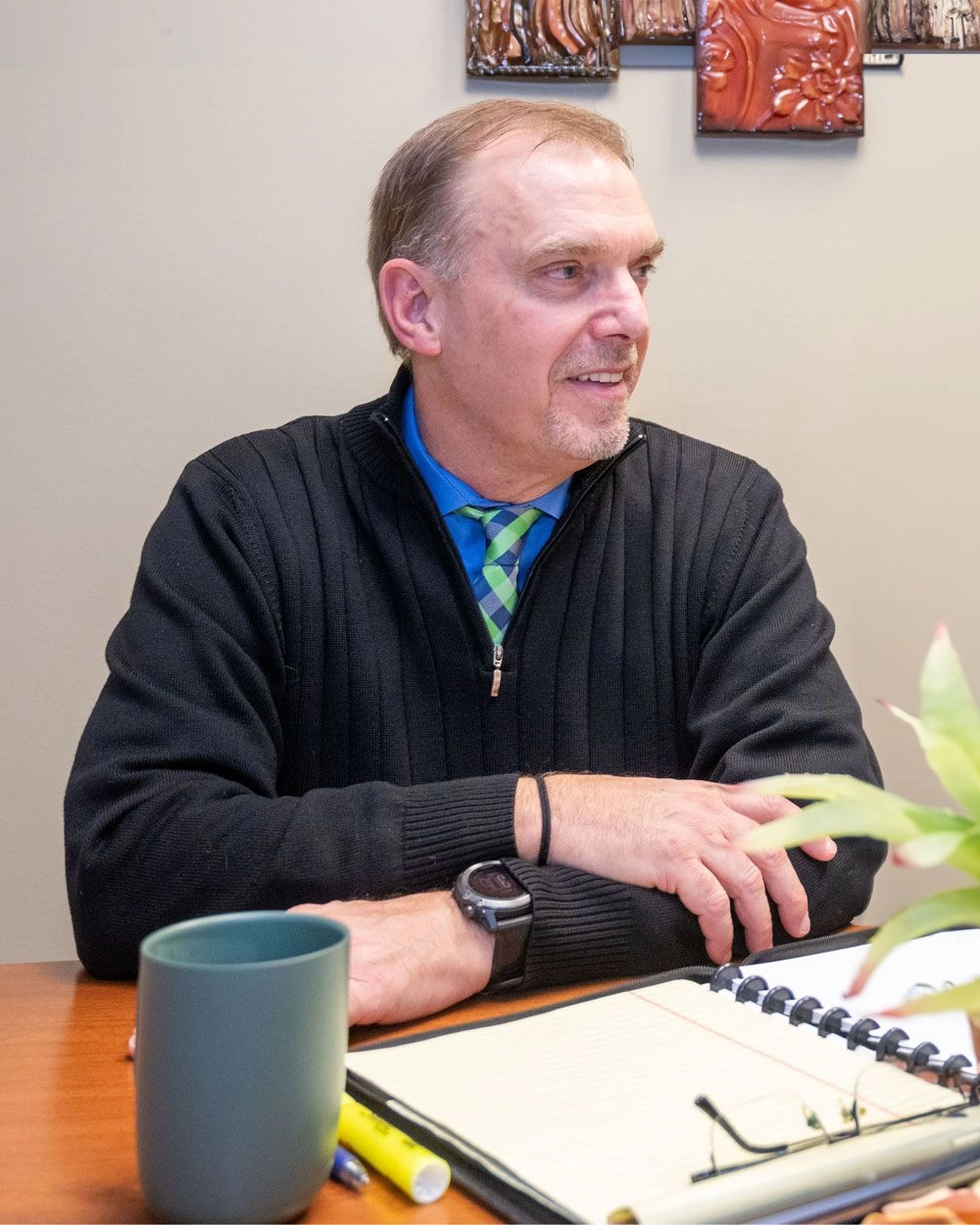 Ed Harper wearing a black sweater and a checkered tie sits at a desk with a notebook, a green mug, a highlighter, and a plant, looking to his right and smiling.