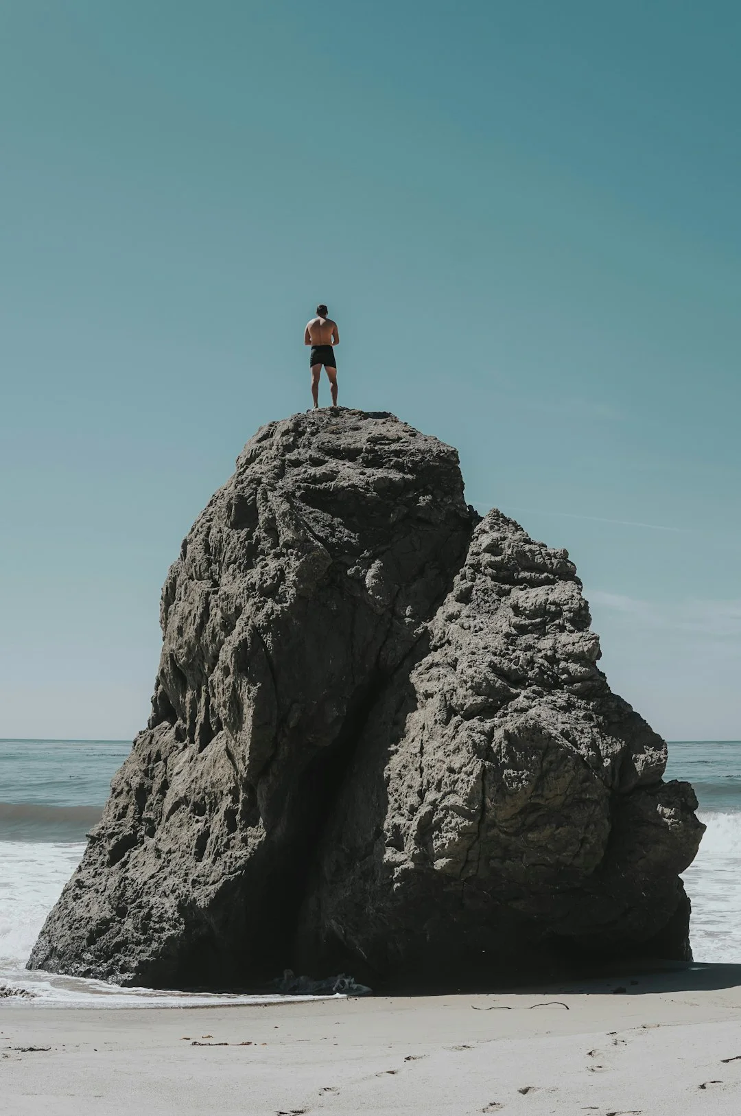 A person stands on a large rock overlooking the ocean under a clear blue sky, with waves gently crashing against the shore in the background.