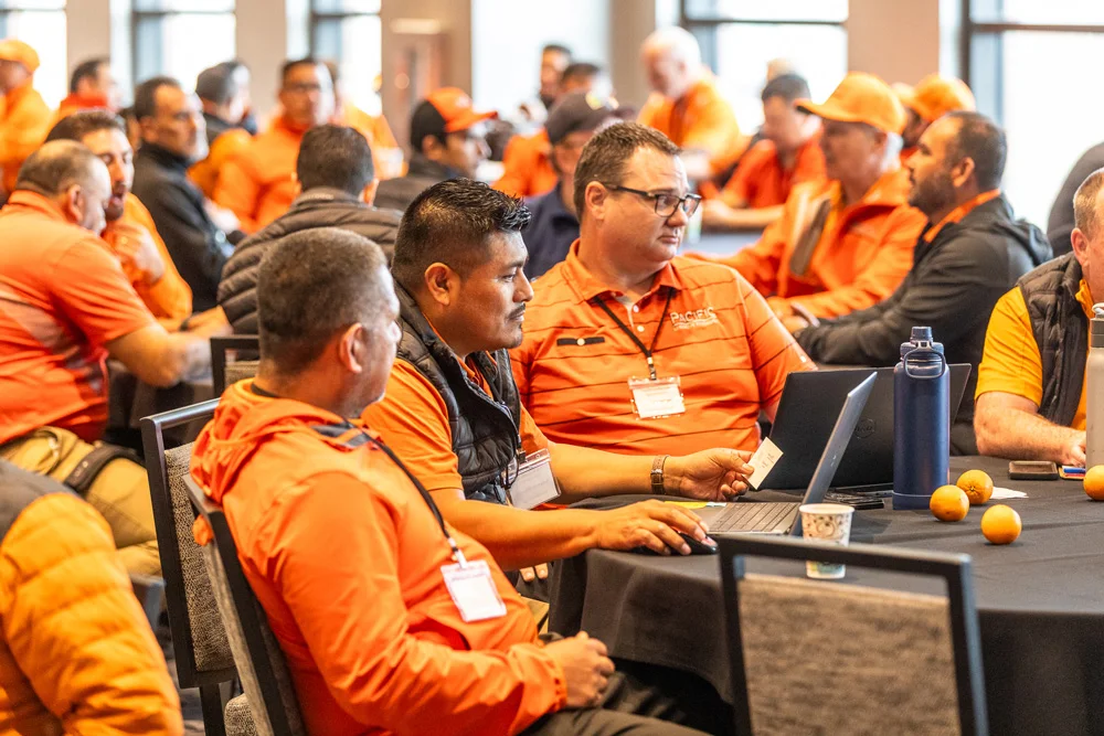 A group of people in orange attire sit at round tables in a conference room, engaging in discussions. Many have laptops open. The room has large windows, allowing natural light.