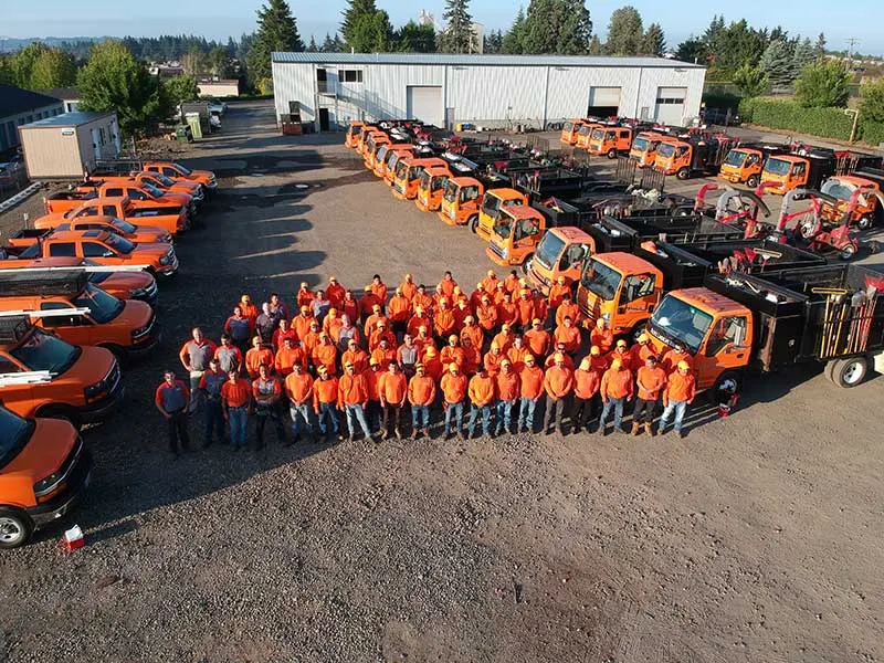 A large group of workers wearing orange safety vests and helmets posing in front of many orange and black utility vehicles in an industrial parking lot with buildings and trees in the background.