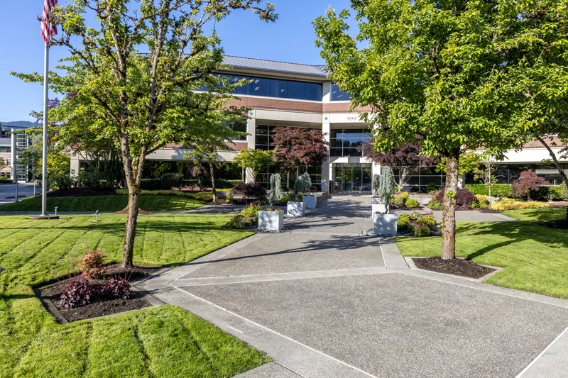 A modern office building with large windows and a landscaped entrance featuring green lawns, trees, and planters on a sunny day. An American flag is visible on the left side.