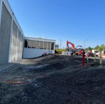 Construction site beside a building with a sloped gravel path. There are construction machines, fencing, and a red fire hydrant visible on a clear, sunny day.