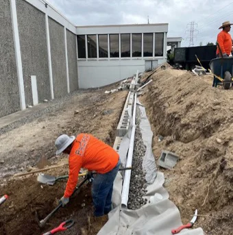 Two construction workers wearing orange shirts and hats are digging a trench near a gray building. One worker is using a shovel while the other stands by a wheelbarrow.