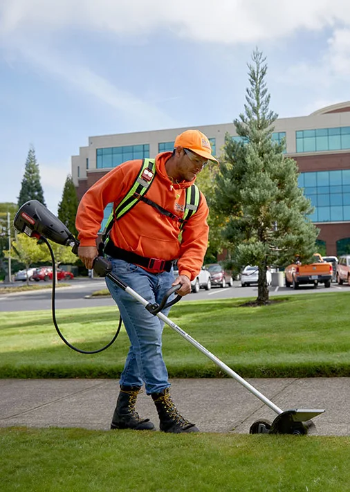 A landscaper in an orange hoodie and cap uses a power edger to trim the grass along a sidewalk next to a grassy area, with a backdrop of modern buildings and trees.