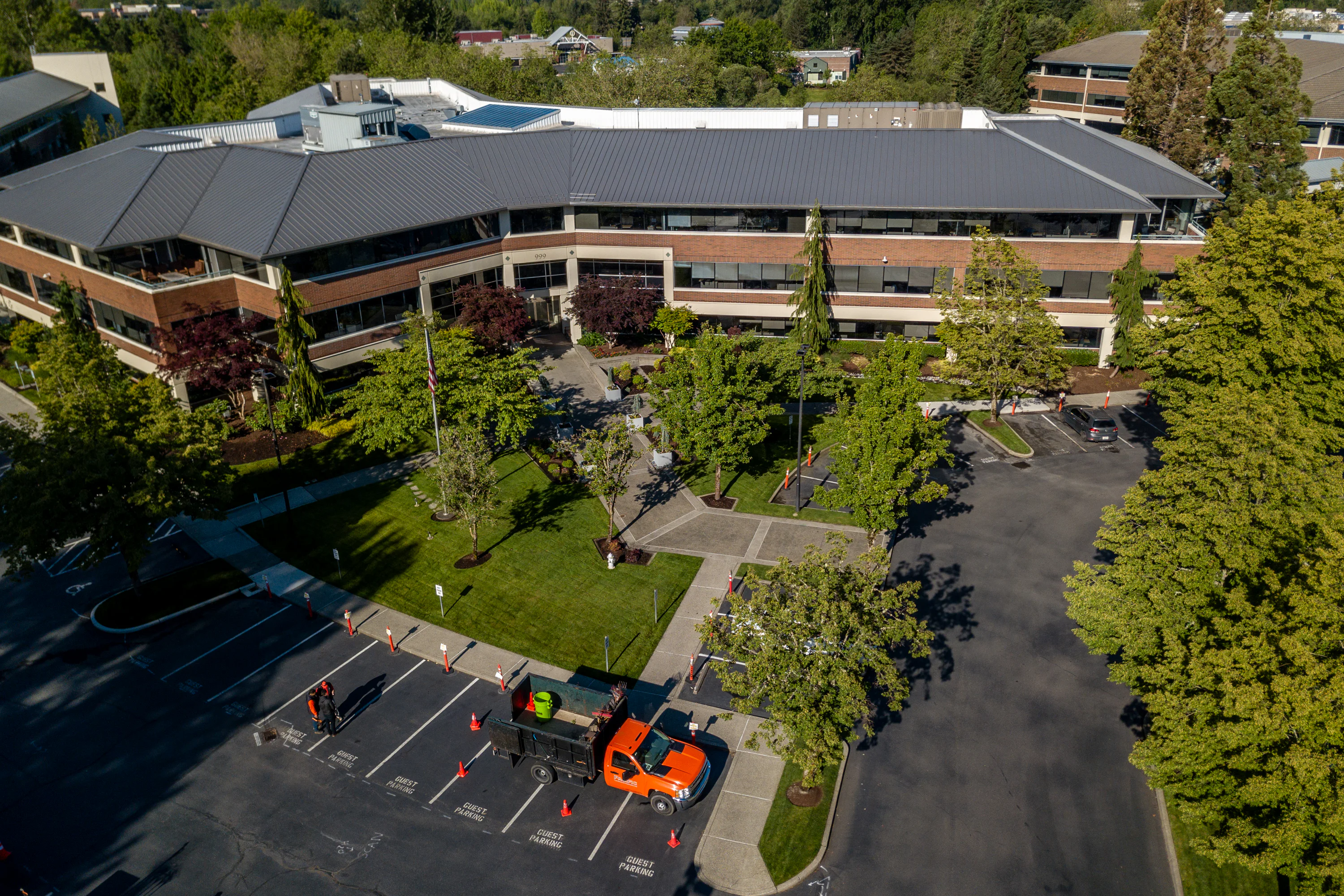 Aerial view of a two-story office building with a dark gray roof, surrounded by trees and well-manicured lawns. An orange truck is parked below, near guest parking spots with traffic cones.