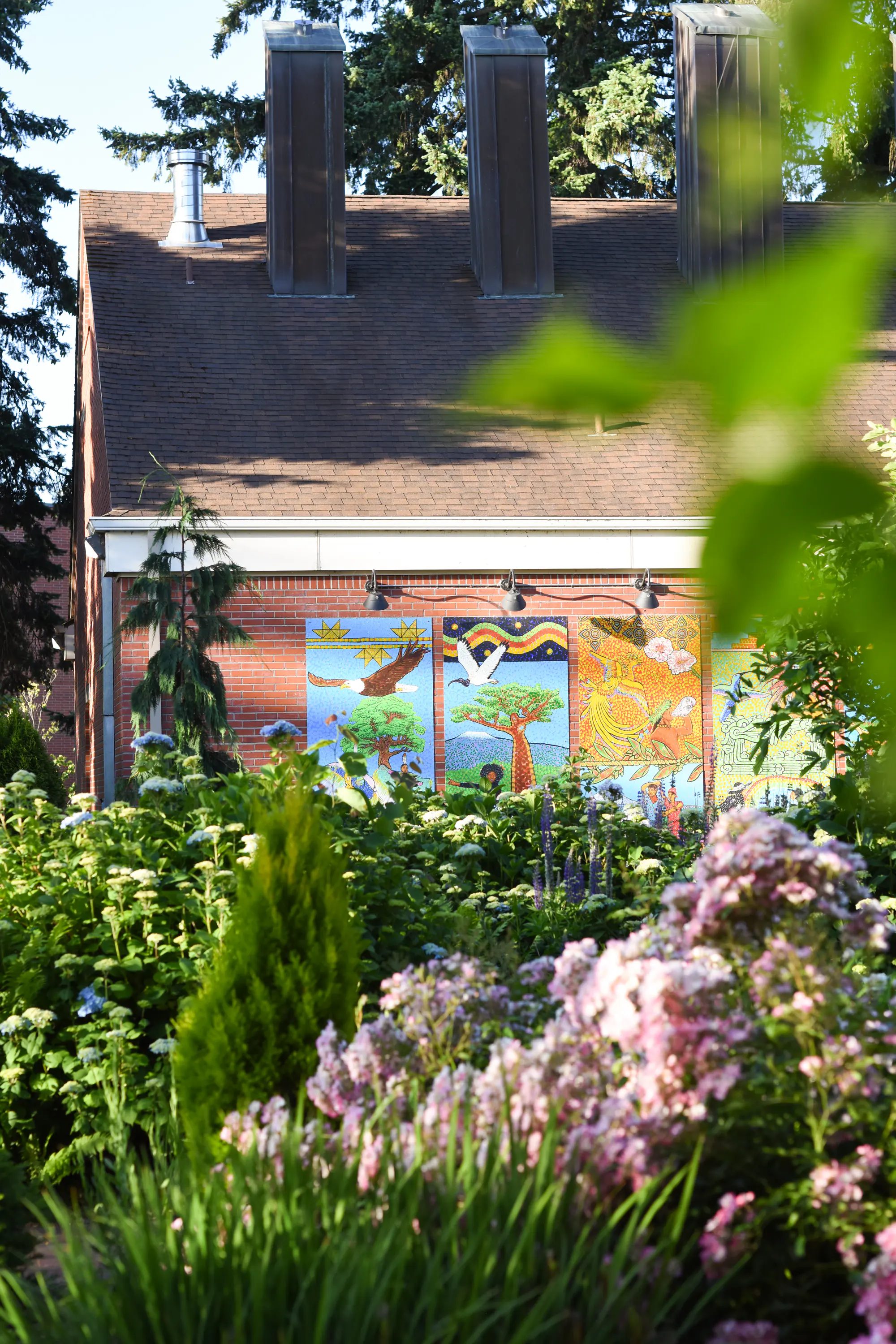 Brick building with colorful mural panels, shingled roof with tall vents, and a lush flowering garden in front