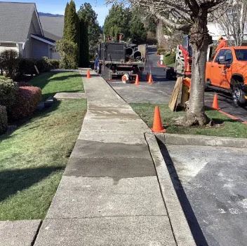 Sidewalk repair in suburban neighborhood with orange cones, trucks, and equipment. Fresh concrete patch visible in the middle of the sidewalk, clear, sunny day with green lawns around.