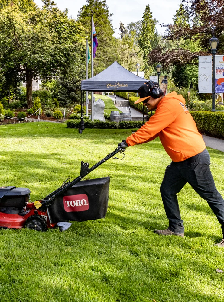 A person in an orange shirt and black pants mows the grass with a Toro lawnmower in a park. A gazebo, trees, and flags are in the background. The setting appears well-maintained and lush.