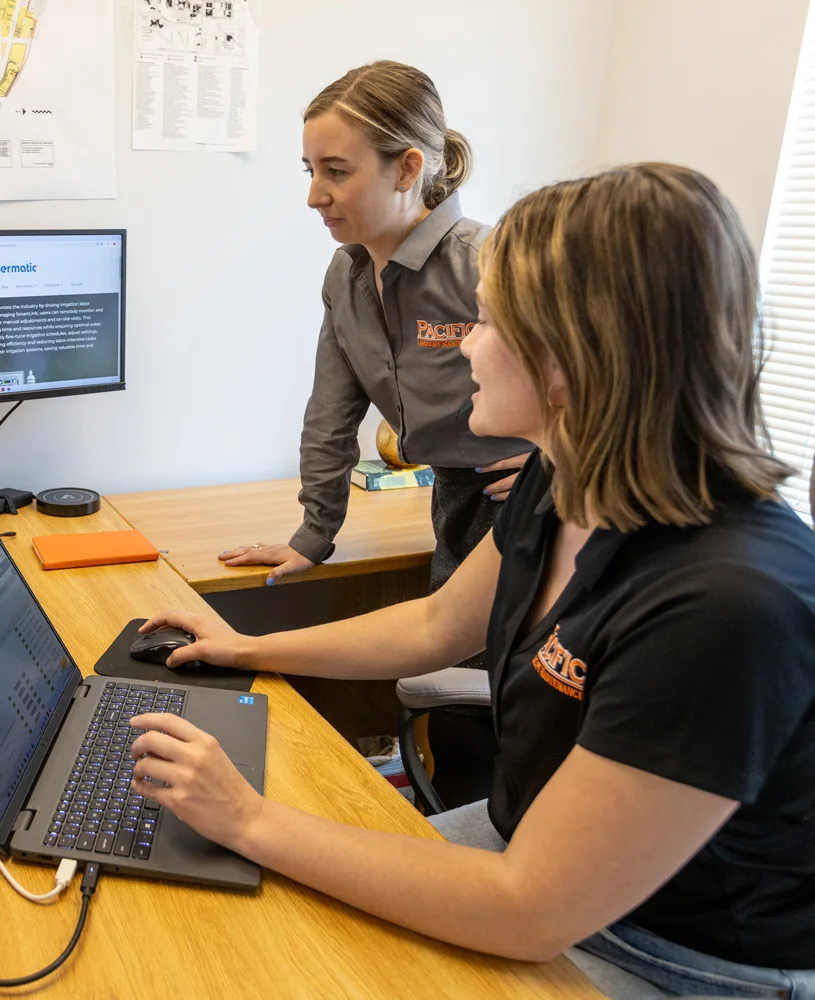 Two women working on a project at a desk, with one using a laptop and the other standing and looking at the screen. Both are wearing shirts with Pacific embroidered.