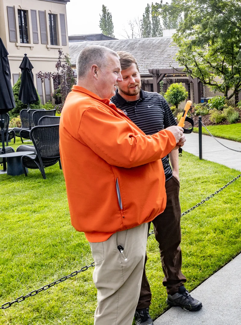 Two men standing on a lawn, looking at something the man in an orange jacket is holding. Black patio furniture and umbrellas are in the background, with trees and a building nearby.