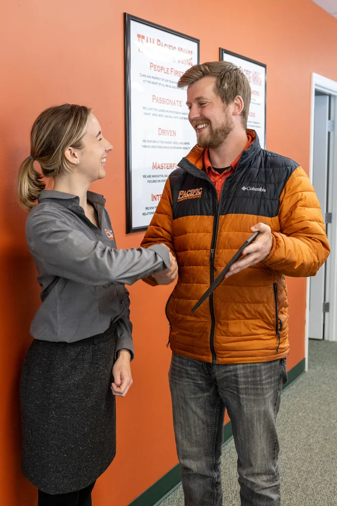 Two people smiling and shaking hands in an office setting with an orange wall. The person on the left wears a grey shirt, and the person on the right wears an orange jacket while holding a tablet.