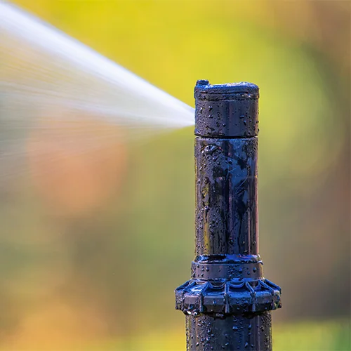 Close-up of a garden sprinkler head spraying water diagonally against a blurred background with green and yellow hues. The sprinkler is wet and appears to be in use.