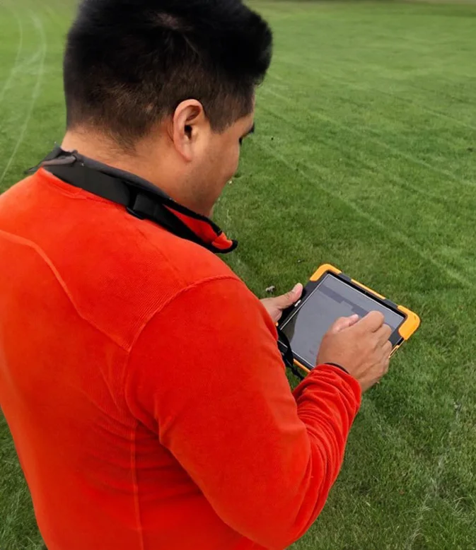 A person in a bright orange shirt uses a tablet while standing on a green lawn, with a black strap around their neck.