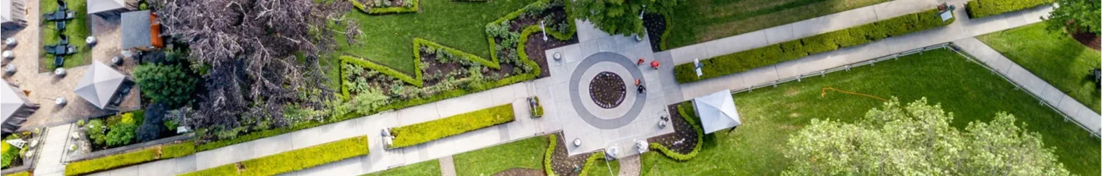 Aerial view of a well-manicured park with winding pathways, benches, and a central circular plaza. Green lawns, hedge designs, large trees, and parking lot with cars are visible.