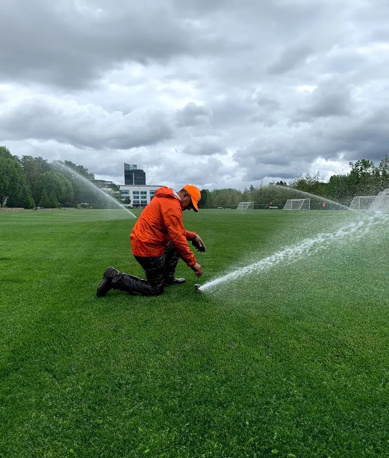 A person in an orange jacket and cap kneels on a grassy field adjusting a water sprinkler system, with cloudy skies and buildings in the background.