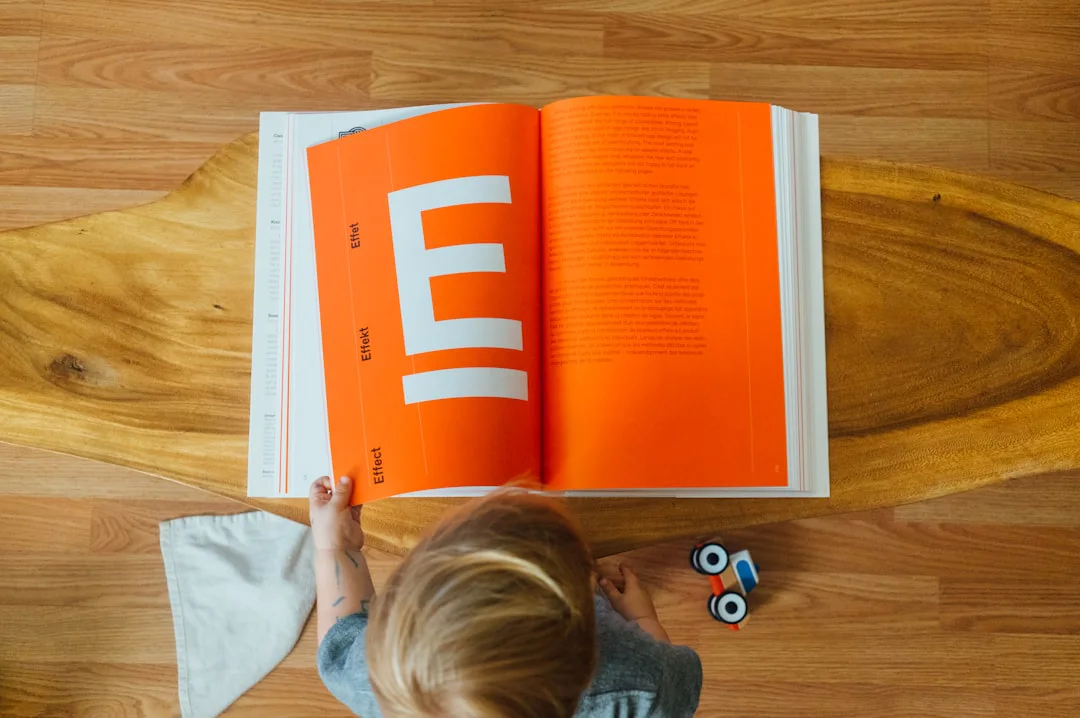 Child viewed from above stands at a wooden table looking at a large open book with bright orange pages. A toy car and white cloth lie on the wooden floor beside the child.