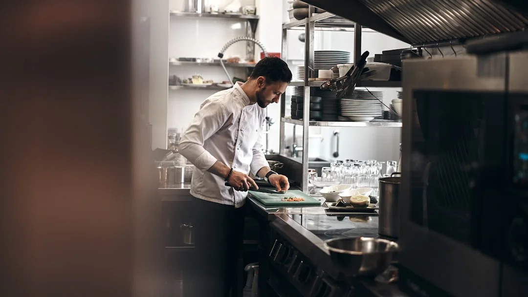 Chef in a professional kitchen chopping ingredients on a green cutting board. Shelves with plates and kitchen tools are visible in the background, along with countertops and appliances.