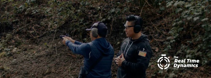 Two men at an outdoor shooting range, one aiming a handgun while an instructor watches; both wear hearing protection